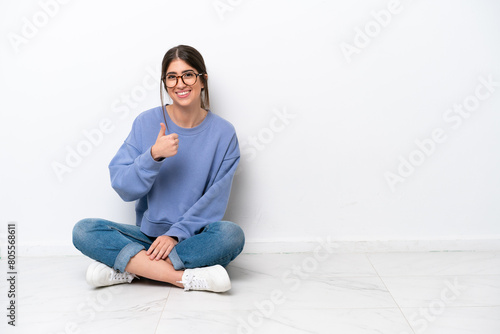 Young caucasian woman sitting on the floor isolated on white background giving a thumbs up gesture