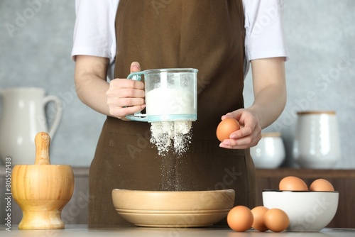 Woman sieving flour into bowl at table in kitchen, closeup