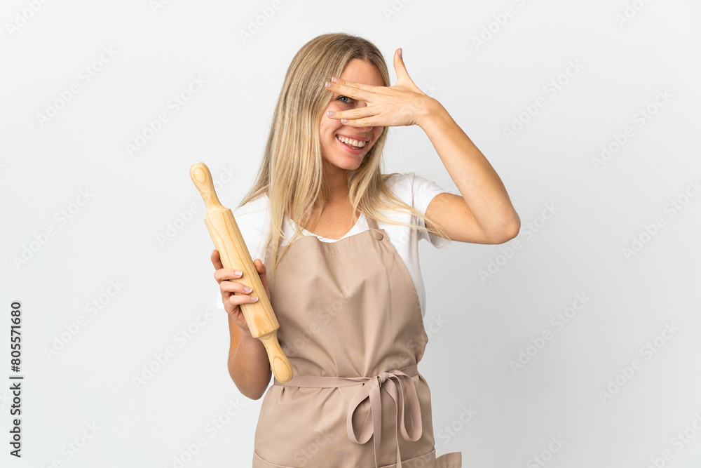 Young woman holding a rolling pin isolated on white background covering eyes by hands and smiling