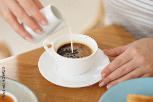 Breakfast. Woman pouring cream into cup of coffee at wooden table, closeup