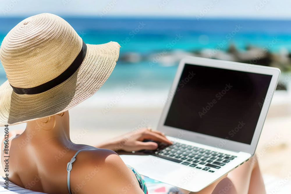 Fototapeta premium Young woman in hat and bikini using laptop on the beach, online business