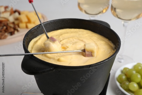 Dipping ham into fondue pot with tasty melted cheese at white table, closeup