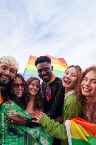 Happy vertical portrait of a group of people celebrating Pride Festival Day with rainbow flags. LGBT community concept. Men and women hugging looking at camera smiling outdoors. Copy space.