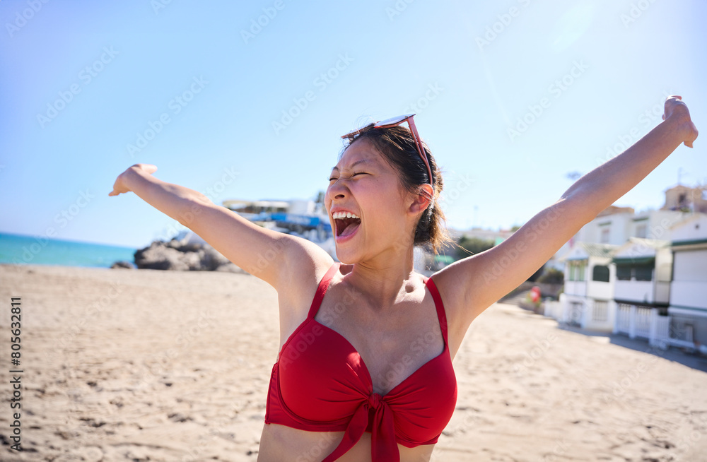 Portrait of young Asian Chinese woman in red bikini with big smile while raising arms on the beach. Happy and cheerful girl on summer holidays. Journeys of generation Z people Portrait of young Asian Chinese woman in red bikini with big smile while raising arms on the beach. Happy and cheerful girl on summer holidays. Journeys of generation Z people