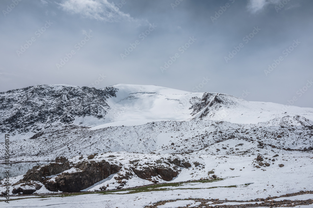 Dramatic cold alpine snow-white landscape with snow-capped peak of dome shape under cloudy sky. Snow-covered rocky mountain with snowy dome shaped top. Pure white snow and stones in high mountains.