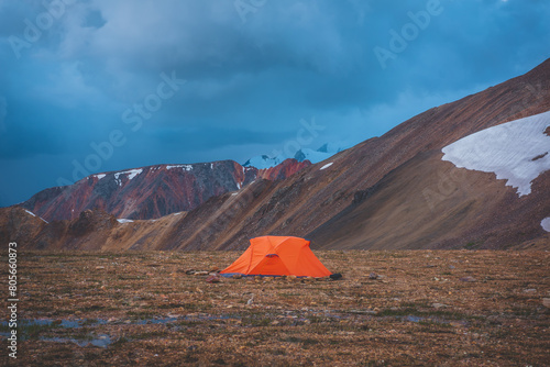 Vivid orange tent on high mountain pass among colorful large mountains with view to great snowy peak in rain in bad weather. Scenic landscape with tent on stony hill among rocks with snow in overcast.