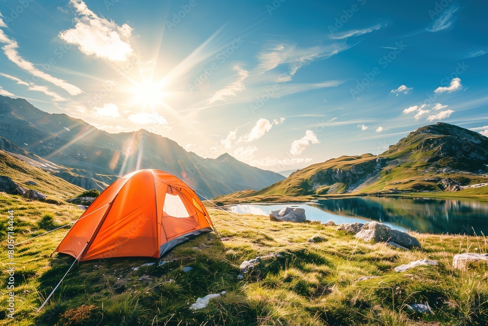Beautiful summer landscape with orange tent on the grass in mountains against blue sky and sunlight, lake at sunrise.