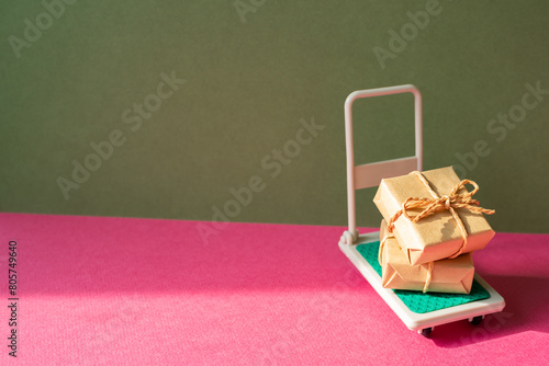 Trolley cart with gift boxes on pink and khaki green background. Parcel Delivery Concept