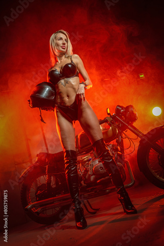 Young sexy girl in the black leather underwear posing near the retro style motorcycle in the red smoke background.