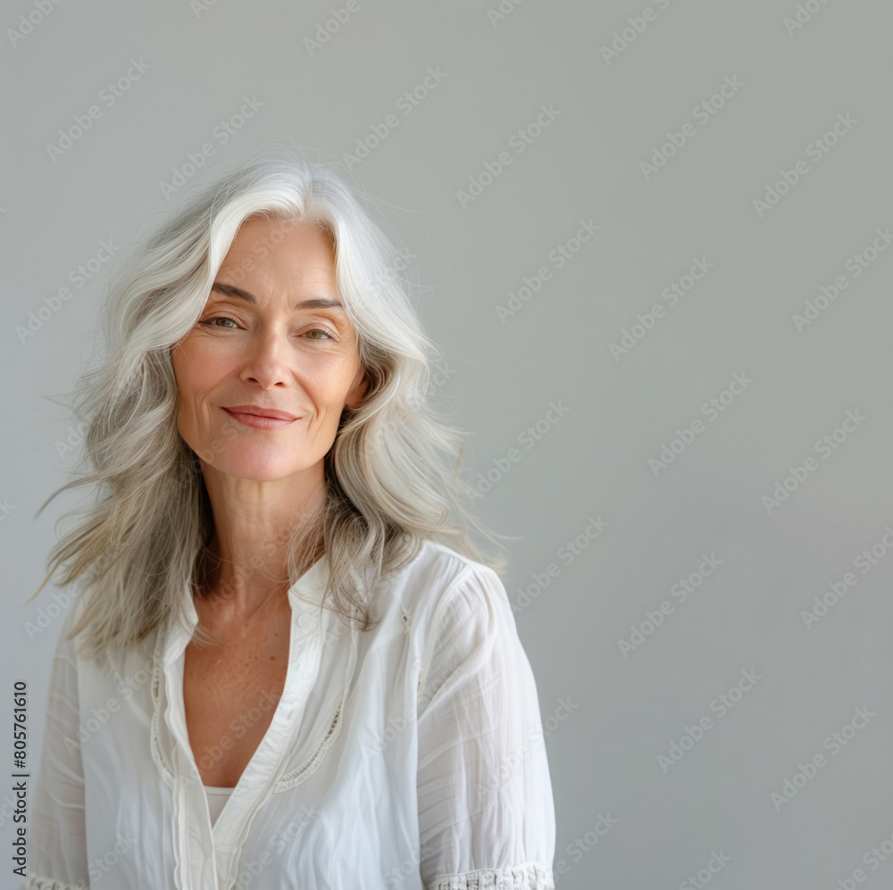 An elegant middle-aged woman with silver hair, wearing a white blouse, poses for a photo shoot against a light grey background, smiling and looking at the camera.