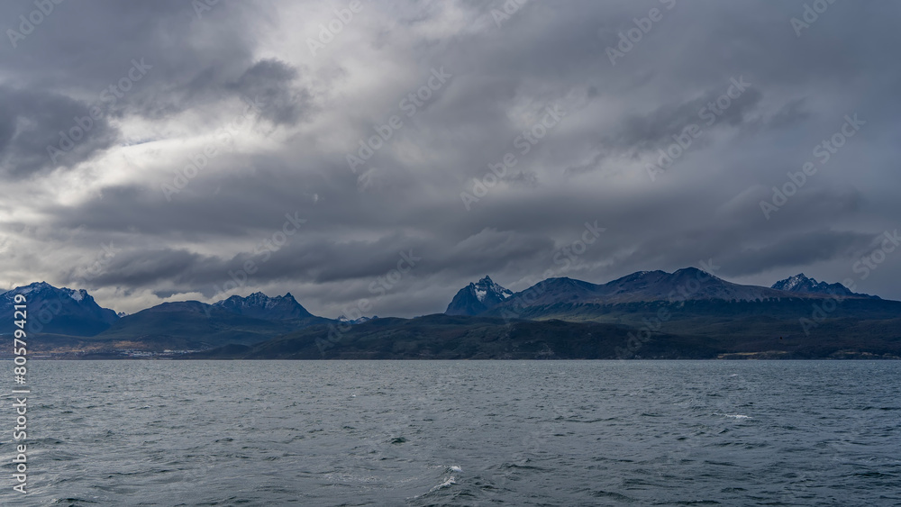 The beautiful snow-capped Andean Martial mountain range is visible from the Beagle Channel. The city of Ushuaia is at the foot, on the ocean. Ripples on the water. Clouds in the sky. Argentina. 