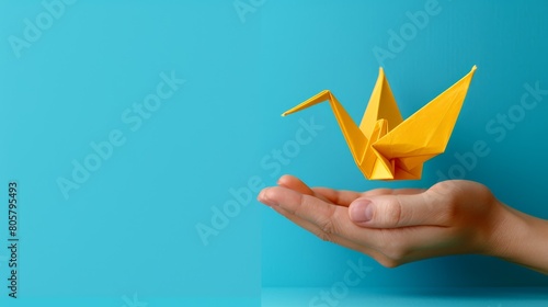   Person's hand holds a yellow origami crane against a blue background Another hand also holds a similar yellow origami crane photo