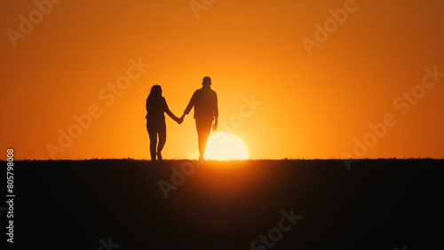 A young couple walks across a field towards the setting sun, holding hands