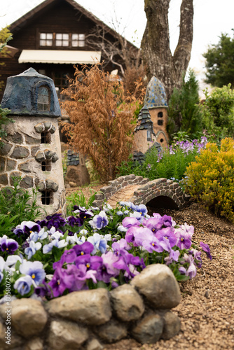 Artistic garden featuring miniature stone house vibrant purple pansies and rustic bridges set against a traditional cabin and tower building vintage