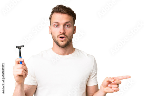 Young caucasian man shaving his beard over isolated chroma key background surprised and pointing side