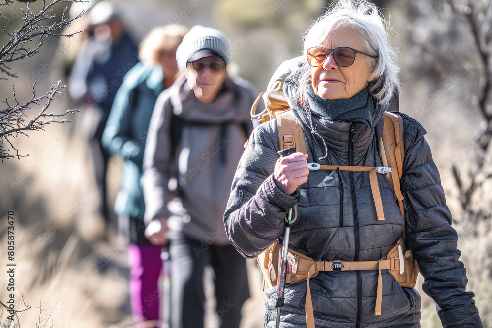 Group of senior smiling gray-haired senior women and men hike a trail in a column through the forest with full gear