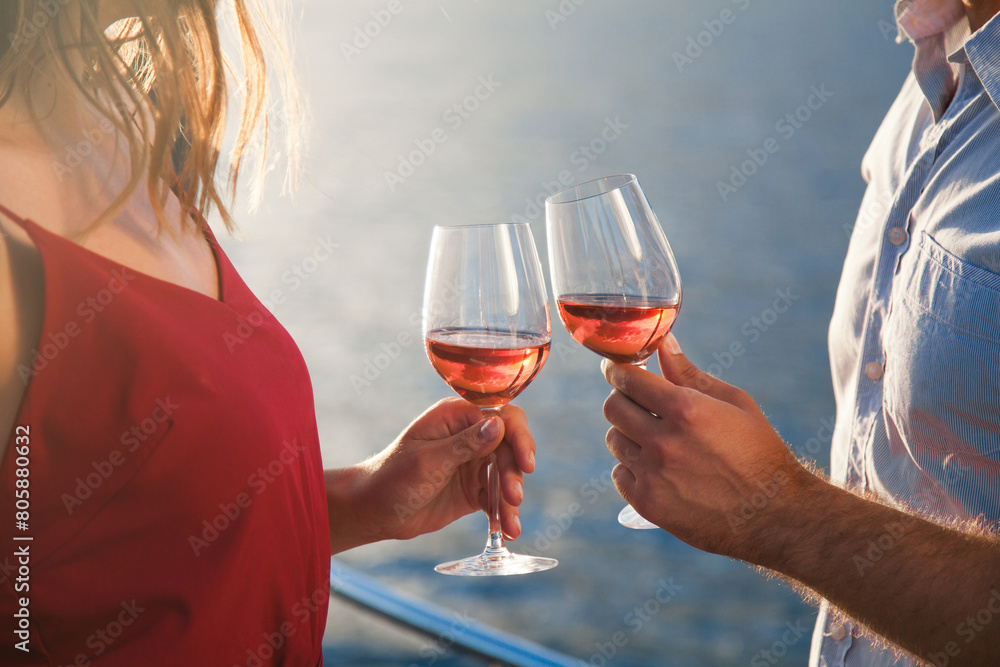 Drinking wine by sea. Two wineglasses in hands of couple in love at sunset beach. Man and woman traveling on yacht, enjoying summer vacation. Romantic celebration on open-air restaurant terrace Drinking wine by sea. Two wineglasses in hands of couple in love at sunset beach. Man and woman traveling on yacht, enjoying summer vacation. Romantic celebration on open-air restaurant terrace