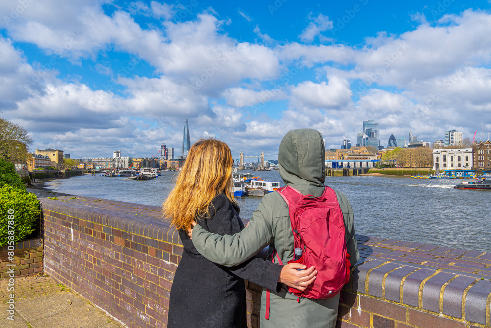 A lesbian couple standing with their backs to each other and embracing, contemplating the Tower Bridge in London and the Shard skyline and the London skyline from the River Thames promenade.