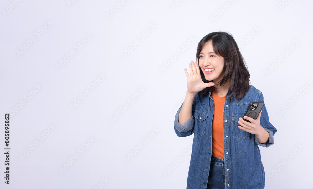 Asian woman wearing orange t shirt and denim jean is posing with a shouting gesture while holding smartphone against a white background. Asian woman wearing orange t shirt and denim jean is posing with a shouting gesture while holding smartphone against a white background.