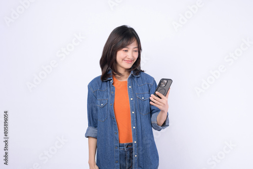 Asian woman wearing an orange shirt and a denim jacket is celebrating the news on mobile phone against a white background.