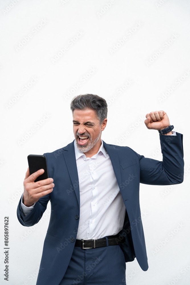 Delighted satisfied business man looking at mobile phone screen gesturing yes winning pose, isolated background. Overjoyed excited businessman in suit make winner gesture reading good news. Vertical Delighted satisfied business man looking at mobile phone screen gesturing yes winning pose, isolated background. Overjoyed excited businessman in suit make winner gesture reading good news. Vertical