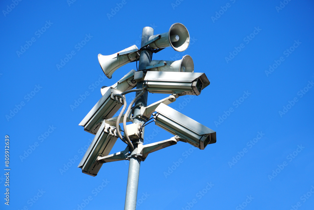 A pole on the street with cameras and loudspeakers mounted on it against a blue sky background. A pole on the street with cameras and loudspeakers mounted on it against a blue sky background.