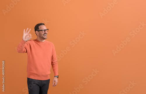 Cheerful male professional laughing and looking away while showing OK sign on orange background