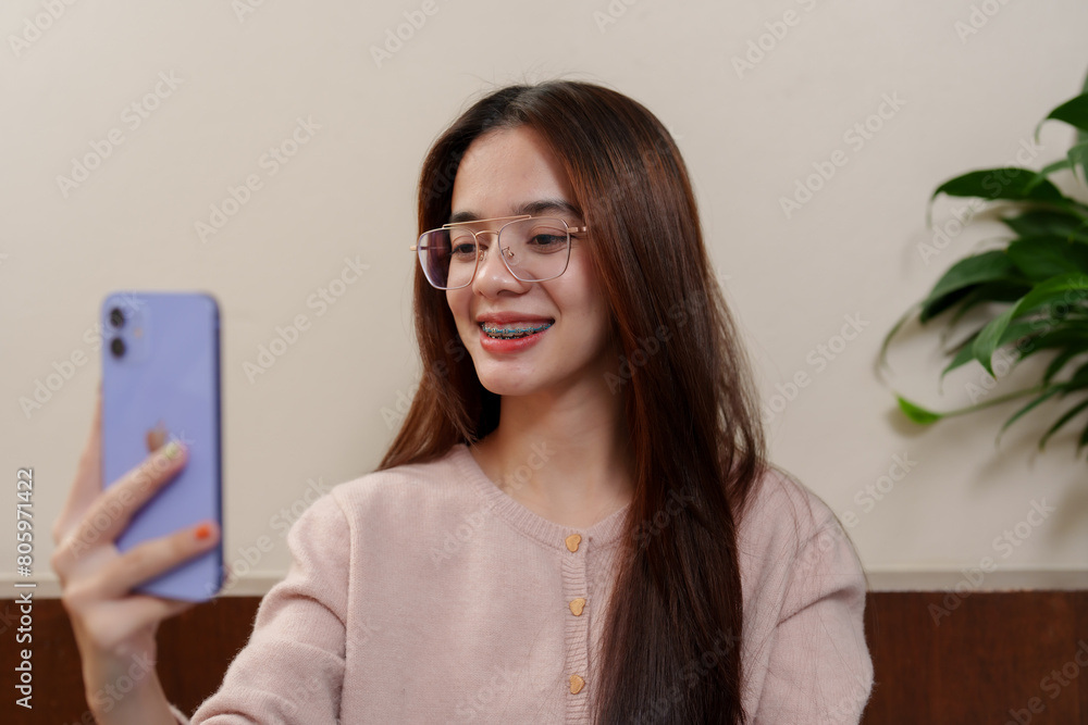 Smiling young person with light brown hair, glasses, braces, taking selfie, potted plant in background, epitomizes relaxed holiday spirit. Holiday happiness for Asian teenagers Smiling young person with light brown hair, glasses, braces, taking selfie, potted plant in background, epitomizes relaxed holiday spirit. Holiday happiness for Asian teenagers