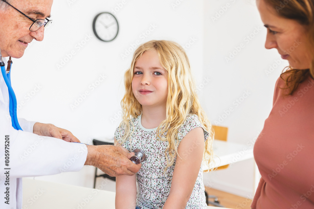 Obraz premium Senior male pediatrician examining sick child with stethoscope at medical room. Little girl visiting doctor with her mother. Health care and medical concept.
