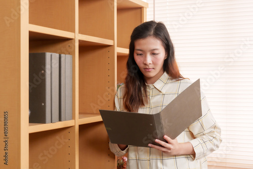 Attentive to details, office worker peruses through paperwork, standing by shelf filled with various folders and office supplies. Office workers are working