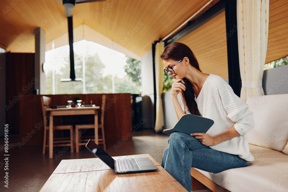 Obraz premium Happy Woman Sitting on Sofa with Laptop and Glasses, Teaching Online from Home A smiling teacher is sitting on a cozy sofa in her living room, working comfortably on her laptop She wears stylish