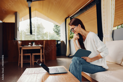 Happy Woman Sitting on Sofa with Laptop and Glasses, Teaching Online from Home A smiling teacher is sitting on a cozy sofa in her living room, working comfortably on her laptop She wears stylish