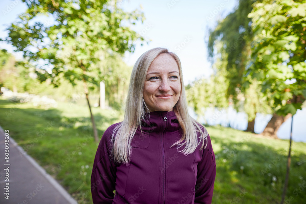 A confident middle-aged woman smiles softly while wearing sportswear in a lush park A confident middle-aged woman smiles softly while wearing sportswear in a lush park