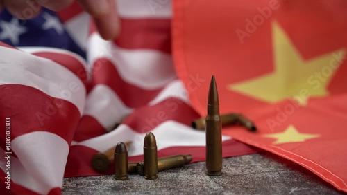 Unrecognizable person's hand pushing a large-caliber bullet placed in line with others, amidst an American and Chinese flag