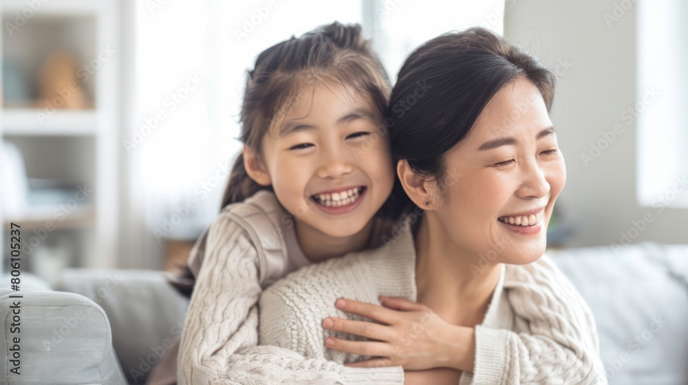 Asian woman sitting on the sofa with her daughter into hugging mom's shoulder, both smiling happy on living room. Asian woman sitting on the sofa with her daughter into hugging mom's shoulder, both smiling happy on living room.