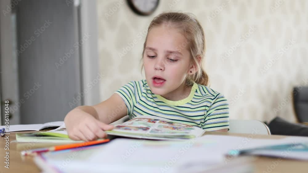A cute little blonde school-age girl is doing her homework sitting at the kitchen table. She reads aloud and does her homework. The concept of online and home education.