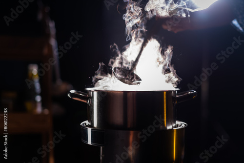 A boiling pot steams up as a chef uses a ladle to stir soup in a cooking pot in a restaurant.