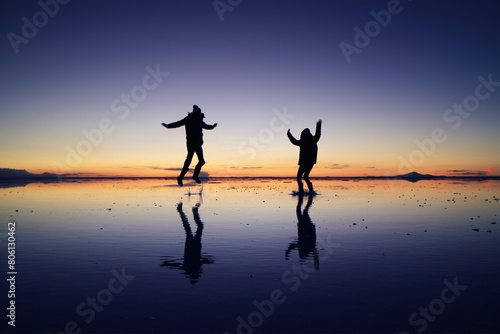 Silhouette of a happy couple jumping on the amazing mirror effect of Uyuni Salt Flats against sunset sky  Bolivia  South America