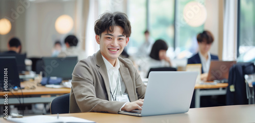 Japanese Business Dynamism: A smiling young businessman, dressed sharply, takes notes on his open laptop at his desk, set against a backdrop of blurred office activity, banner