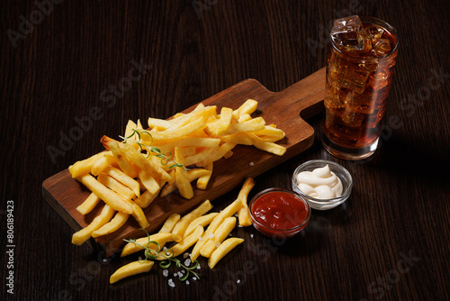 A pile of French fries on a wooden board. Nearby there are gravy boats with mayonnaise and ketchup on a dark wooden table, and cola with ice in a glass glass. Angled view.