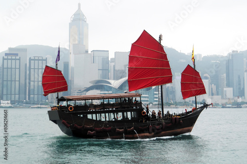 old Chinese boat (junk) with red sails; a group of tourists on a pleasure trip along the Victoria Strait (Hong Kong, China); skyscrapers; morning mist