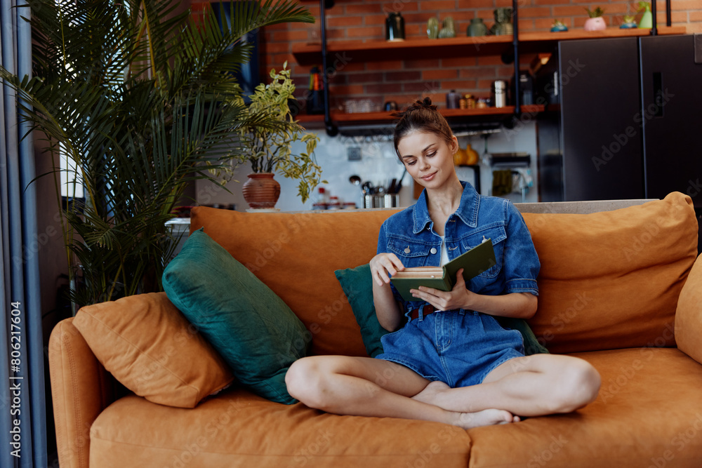 Obraz premium Young woman enjoying leisure time at home, reading a book on orange couch in denim shirt