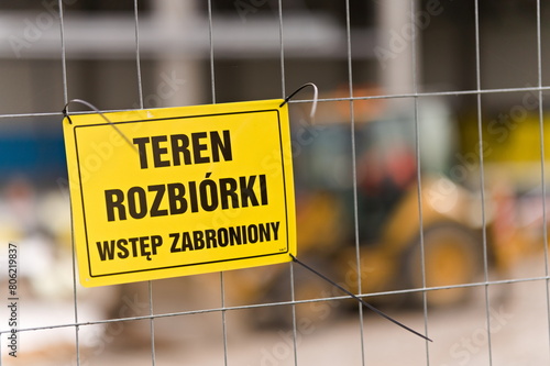 Znak Teren rozbiórki Wstęp zabroniony wiszący na płocie, w tle zarys koparki oraz budynku podczas budowy