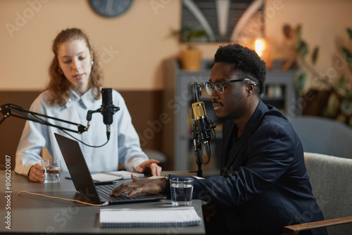 Side view of African American male podcaster in suit and eyeglasses typing on laptop and listening to Caucasian female guest sitting in studio
