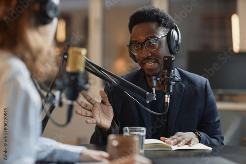 Medium shot of young Black man in eyeglasses and suit answering questions of unrecognizable interviewer during podcast recording