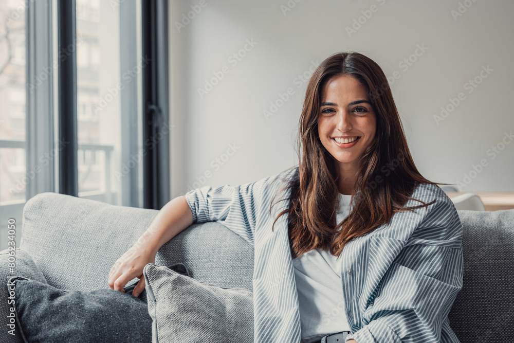 Naklejka premium Happy attractive young adult woman girl sitting on sofa looking at camera in modern cozy home, smiling single 20s lady beautiful face relaxing on couch alone enjoy wellbeing posing for portrait.