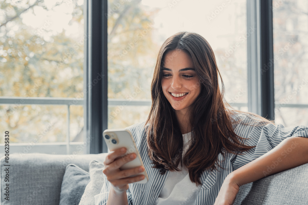 Happy pleasant millennial woman relaxing on comfortable couch, holding smartphone in hands. Smiling young lady chatting in social networks, watching funny videos, using mobile applications at home..