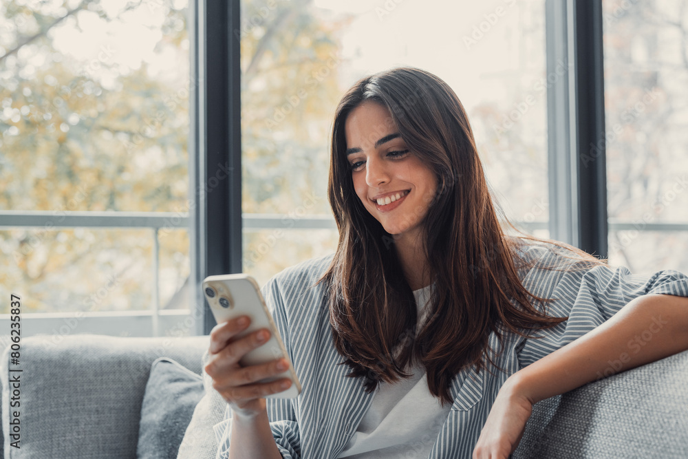 Happy pleasant millennial woman relaxing on comfortable couch, holding smartphone in hands. Smiling young lady chatting in social networks, watching funny videos, using mobile applications at home..