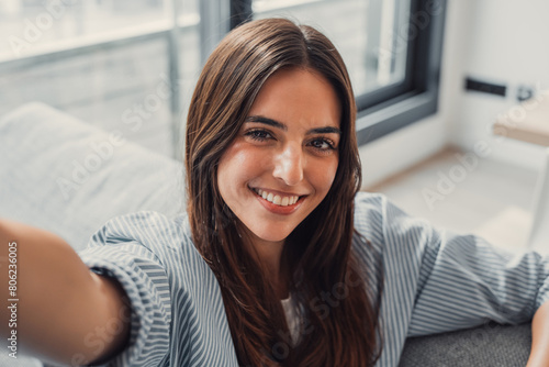 Happy attractive young adult woman housewife sitting on sofa looking at camera in modern cozy home, smiling single 20s lady beautiful face taking a selfie alone enjoy wellbeing posing for portrait.