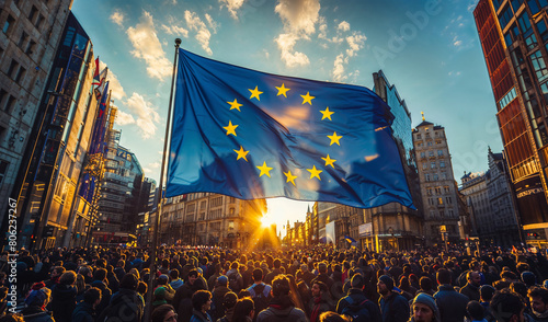 A large gathering of people fills a city square, a European Union flag waving prominently as the sun sets in the background. photo